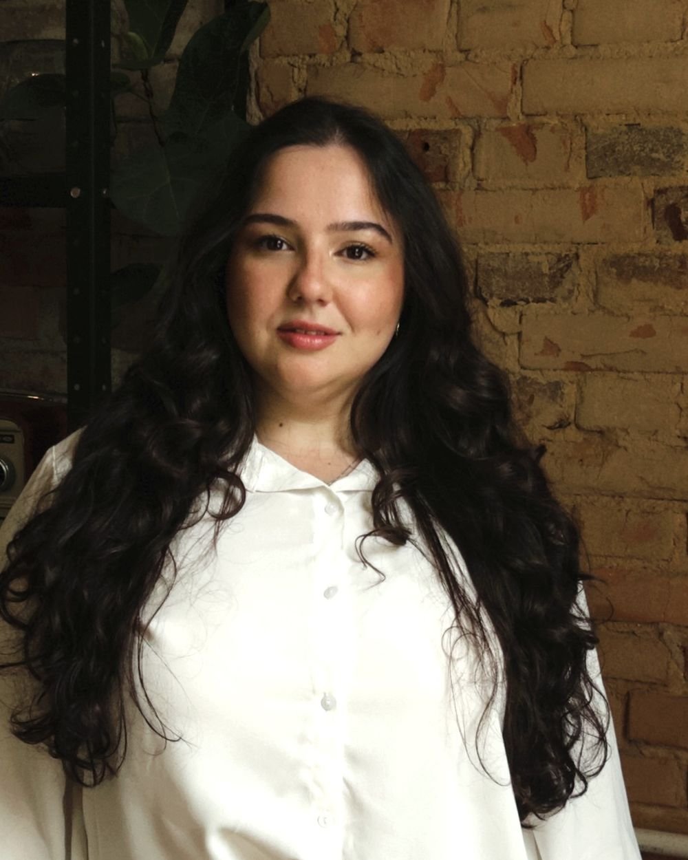 Vivian Andrade em pé, de camisa branca, diante de uma parede de tijolos